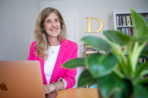 Daphne is shown sitting at her laptop in her office. A plant is in the foreground. She wears a pink jacket and smiles at the camera.