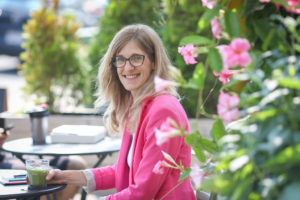 Daphne is shown in an outdoor location sitting amongst greenery and pink flowers. She looks at the camera and smiles. She holds a pen and a green matcha latte rests on the table in front of her.