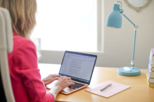 Daphne is shown at her desk, typing on her laptop. A desk laps is nearby, as well as a notebook and pen.