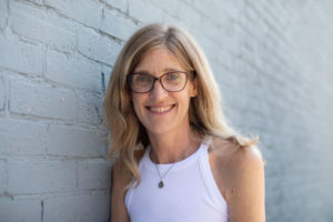 Daphne's face is shown. She stands against a gray brick wall in an outdoor location and smiles at the camera.